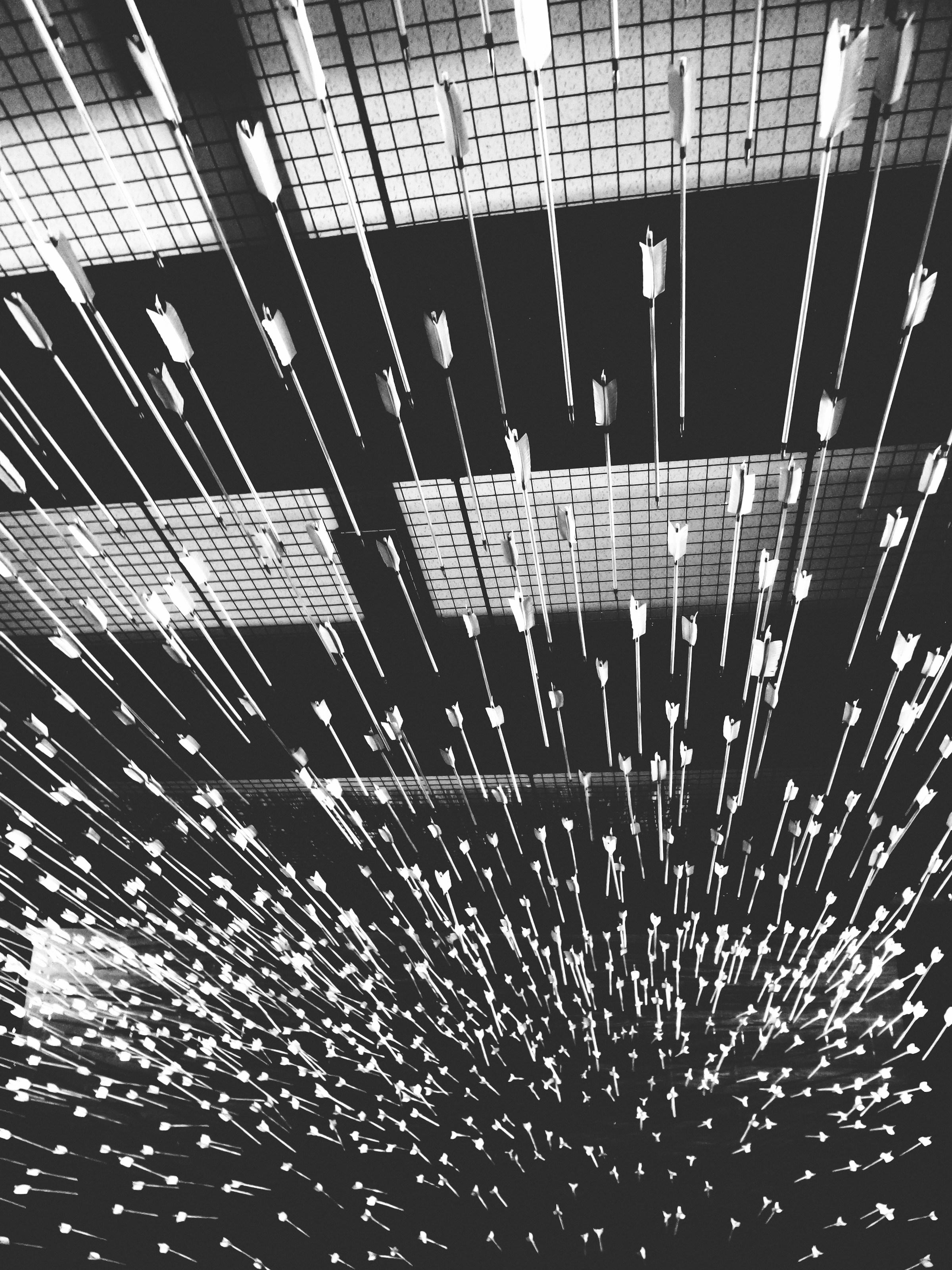 A black and white image inside a swarm of arrows flying in the same direction through a darkened warehouse with dim ceiling lights toward darkness, viewed from inside the swarm with shafts closer to the viewer and arrowpoints away toward the target